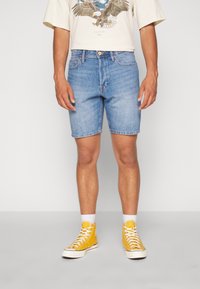 Shorts de mezclilla en azul claro, corte recto, con dobladillo sin terminar. Combinados con una camiseta gráfica color crema y zapatillas altas amarillas. Detalles de costura bien cuidados visibles.