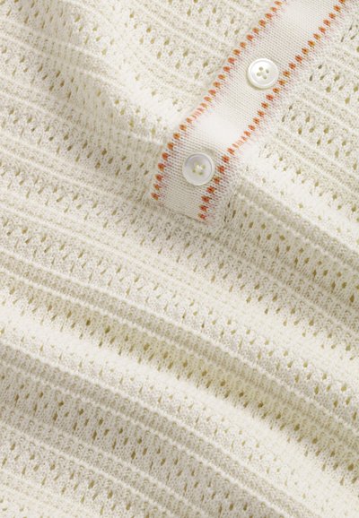 Tessuto lavorato a maglia di colore crema con un motivo testurizzato di punti aperti. Presenta un collo a costine bianco con accenti arancioni e due bottoni.