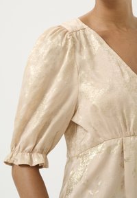 Abito color beige chiaro con finitura luccicante, scollatura a V, maniche corte a sbuffo e dettagli arricciati in vita, con polsini arricciati.