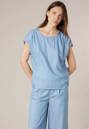 Femme debout, mains dans le dos, portant un haut bleu clair à manches courtes et un pantalon large assorti, sur fond beige.