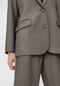 Blazer gris sur mesure avec revers crantés, deux poches avant et deux boutons. Tissu texturé avec un motif subtil, assorti à un pantalon coordonné.