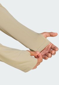 Ruky sťahujúce béžové kompresné návleky s otvormi na palce, ktoré siahajú cez zápästia, na jednoduchom pozadí.