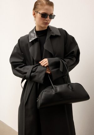 Vrouw in een oversized zwarte jas met glanzende kraag en zwarte zonnebril, die een lange zwarte leren handtas vasthoudt tegen een effen achtergrond.