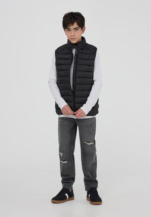 Gilet invernale nero con colletto alto, chiusura con zip e motivo trapuntato, indossato sopra una camicia bianca a maniche lunghe, con jeans grigi strappati e scarpe nere.