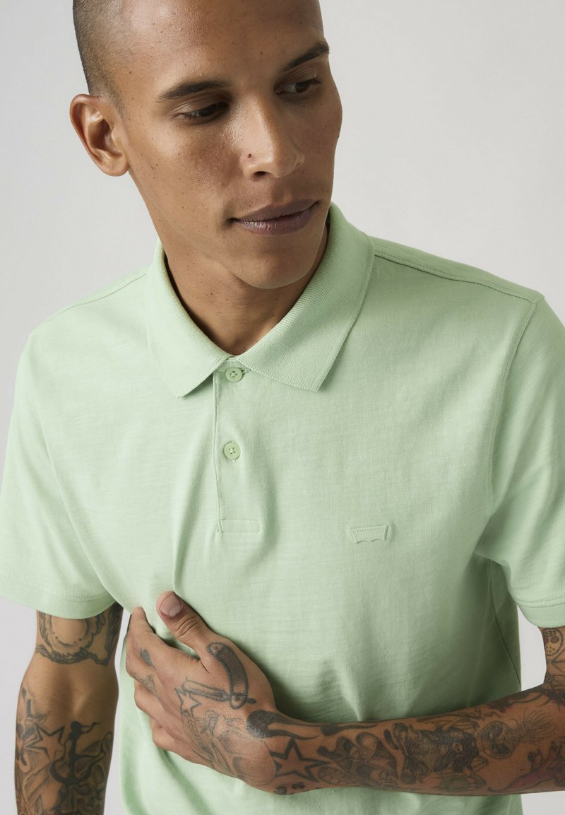 Man met kort haar en tatoeages op beide armen draagt een lichtgroen poloshirt, kijkt omlaag en houdt het shirt dicht bij zijn borst vast.