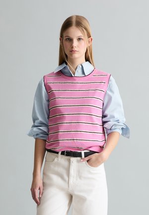 Jonge vrouw met steil blond haar, een roze gestreepte mouwloze trui over een lichtblauw overhemd en witte broek, met haar hand in haar zak.