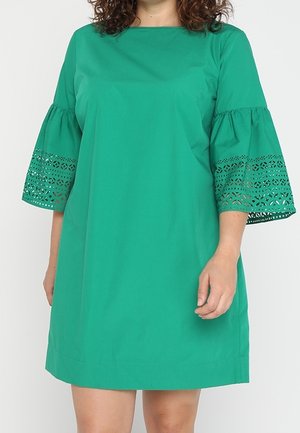 Mujer con un vestido verde hasta la rodilla con amplias mangas de tres cuartos con detalle de encaje de ojales.