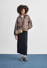 Καφέ ζακέτα με φερμουάρ και σχέδιο λεοπάρ με μαύρες ρίγες, μαύρη μίντι φούστα με πλευρική σχισμή, γκρι αθλητικά παπούτσια και μπεζ τσάντα χειρός με λεπτομέρεια λογότυπου.