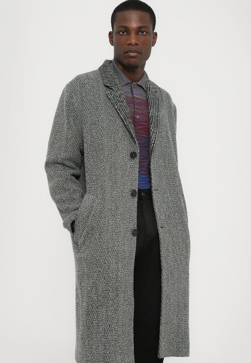 Grijze visgraatwol coat met notch-kraag, twee voorzakken en knopen aan de voorkant, gedragen over een meerkleurig gestreept shirt.