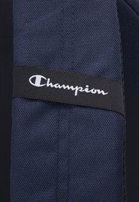 Sac en tissu bleu marine avec une surface texturée, comportant une étiquette noire avec le logo "Champion" blanc cousu sur le côté.