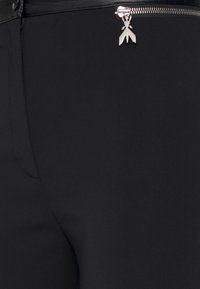 Pantalones negros ajustados con un diseño aerodinámico, que incluyen un cierre de cremallera metálica y un acento decorativo plateado cerca de la cremallera.