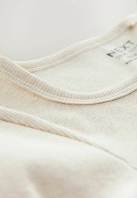 Cremfarbener gerippter Stoff mit einem runden Ausschnitt und feiner Textur, mit sichtbarem Markenlabel. Nahaufnahme.