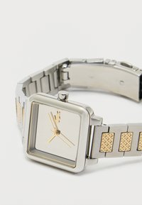 Hõbedane roostevabast terasest kell ruudukujulise näo, kuldsete detailide ja tekstureeritud rihmaga, millel on ristkülikukujulised mustriga lingid.