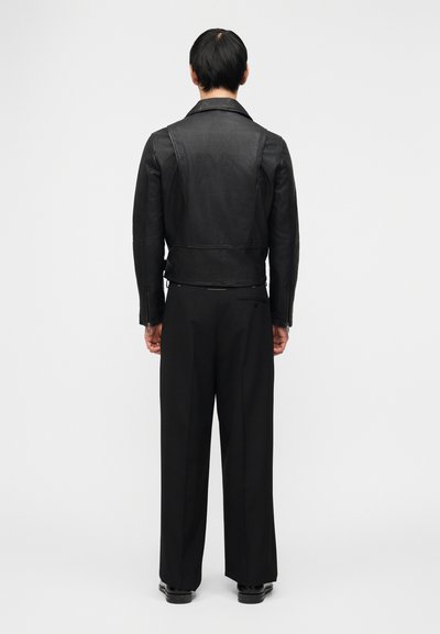 Άνδρας που στέκεται με την πλάτη, φοράει μαύρ ο δερμάτινο μπουφάν, μαύρο παντελόνι με φαρδιά μπατζάκια και μαύρα παπούτσια, μπροστά σε απλό λευκό φόντο.