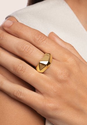 Goldring mit einem glatten, geometrischen Design, das eine auffällige, gebogene Oberfläche aufweist, auf einer Hand vor einem hellen Stoffhintergrund.