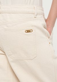 Beige denim pants featuring a back pocket with a gold-tone logo accent and visible stitching. The fabric has a smooth, textured finish.