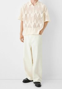 Camicia corta color crema con motivo a zigzag, caratterizzata da un design con bottoni sul davanti e una vestibilità ampia. Indossata con pantaloni a vita alta e gamba larga.