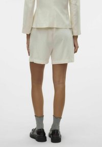 Pantaloni corti beige chiaro con vestibilità rilassata, realizzati in tessuto morbido, mostrati da dietro. Abbinati a calzini grigi e scarpe nere.