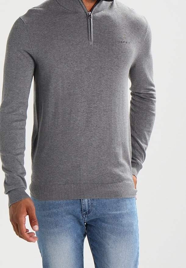 Mann trägt einen grauen Viertelzip-Pullover mit gerippten Bündchen und Saum, kombiniert mit hellblauen Jeans, und steht vor einem schlichten Hintergrund.