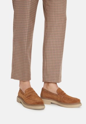 Brązowe mokasyny zamszowe z grubymi beżowymi podeszwami noszone z przyciętymi brązowo-beżowymi spodniami w kratę pepitkę.