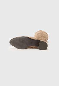 Bottine en suédine beige avec un bout arrondi et une semelle texturée. Le talon est court et légèrement évasé, avec des coutures verticales le long des côtés.