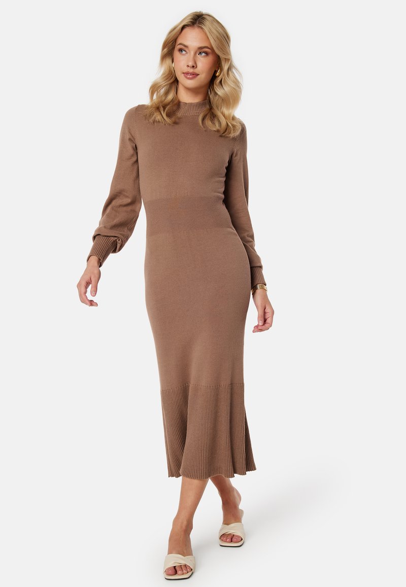 Robe tricotée marron avec un ourlet et des manches côtelés. Elle présente un corsage ajusté, un col montant et une partie inférieure évasée, accompagnée de sandales de couleur claire.