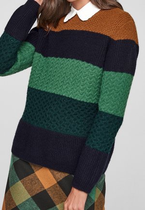 Vrouw draagt een trui met kleurvlakken en textuur in bruin, marineblauw en groen over een wit overhemd met kraag, gecombineerd met een geruite rok in bruin en groen.