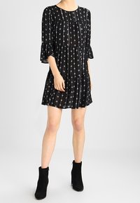 Schwarzes Kleid mit ausladenden Ärmeln, das mit einem vertikalen Muster aus weißen und silbernen geometrischen Formen versehen ist. Kurze, lockere Passform. Kombiniert mit schwarzen Ankle Boots.