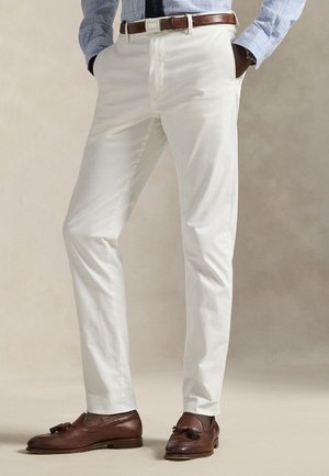 Homme portant un pantalon blanc slim, une ceinture en cuir marron, une chemise à carreaux bleue et des mocassins en cuir marron à pampilles, debout les mains dans les poches.