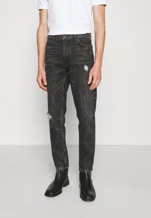 Hombre con jeans negros desgastados, botas negras hasta el tobillo y una camisa blanca de manga corta, de pie contra un fondo blanco liso.