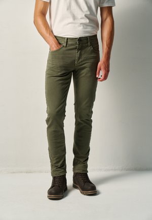 Hombre con pantalones verde oliva, camiseta blanca y botas oscuras con cordones, de pie con una mano en el bolsillo.