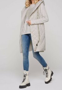 Ljust grått quiltat lång väst med huva, med sidofickor och ett dekorativt band. Matchas med blå skinny jeans och vita snörboots.
