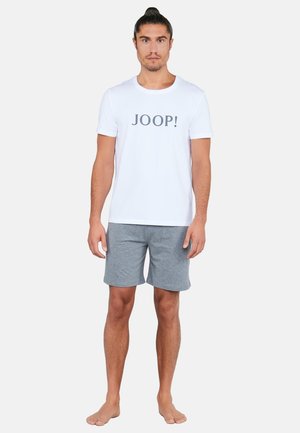 Mann steht barfuß, trägt ein weißes JOOP! T-Shirt und graue Shorts vor einem schlichten hellen Hintergrund.