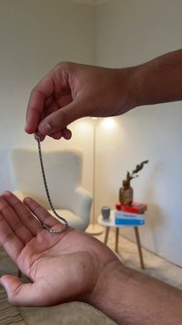 Corrente de metal prateado segurada entre as mãos, apresentando segmentos ligados e um fecho circular. Fundo suave com móveis e uma planta.