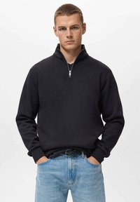 Zwarte zip-up trui met een hoge kraag, lange mouwen en geribde manchetten, in combinatie met lichtblauwe jeans. Soepele stof, minimalistisch ontwerp.