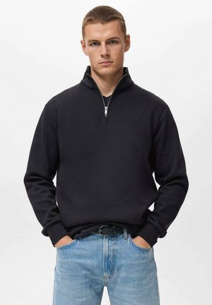 Pulover negru cu fermoar, cu guler înalt, mâneci lungi și mansete cu elastic, asociat cu blugi de un albastru deschis. Material neted, design minimalist.