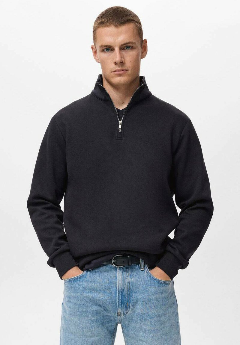 Zwarte zip-up trui met een hoge kraag, lange mouwen en geribde manchetten, in combinatie met lichtblauwe jeans. Soepele stof, minimalistisch ontwerp.