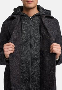 Abrigo oscuro de espiga con un acabado texturizado, que presenta un gran cuello, detalles con botones y una sudadera gris con capucha y cremallera oculta debajo.