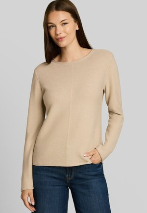 Junge Frau mit glatten braunen Haaren, die ein beiges Langarmshirt und dunkelblaue Jeans trägt, stehend mit einer Hand in der Tasche.
