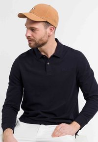 Mornarsko dolg rokav polo majico z zapenjanjem, nosi svetlo bež kapo. Ima subtilen logotip na prsih in rebraste manšete.