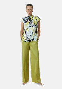 Bloem blouse in lichtblauw, zwart en geel; korte mouwen. Groene wijde broek; gladde textuur. Open teen sandalen met witte banden.