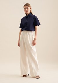 Granatowa koszula z krótkim rękawem zapinana na guziki, zestawiona z białymi, wysokim talii szerokimi spodniami i czarnymi sandałami na obcasie, na neutralnym tle.