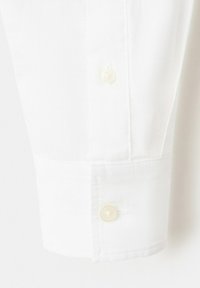 Poignet de chemise blanche à manches longues avec deux boutons écrus et des détails de couture visibles sur un fond uni.