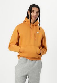 Sweatshirt de algodão laranja com um bolso canguru e o logo branco da Nike no peito, combinado com calças de moletom cinzentas.