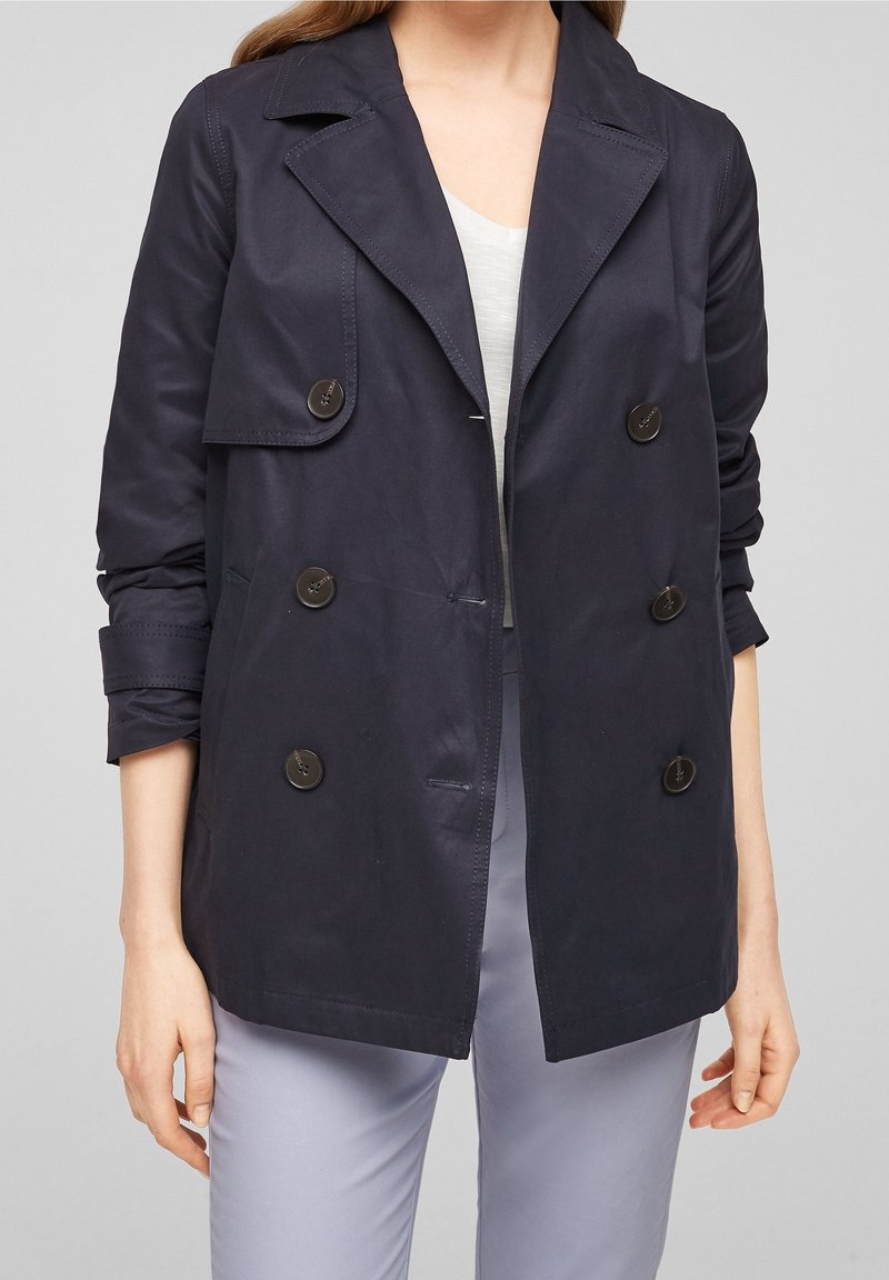 Granatowy, dwurzędowy płaszcz trench z wywijanym kołnierzem, z dużymi guzikami i podwiniętymi rękawami, wykonany z gładkiego materiału.