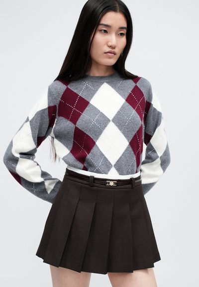 Sivý sveter s červeným a bielym argyle vzorom a trblietavými akcentmi, skombinovaný s tmavohnedou skladanou sukňou a úzkym bielym opaskom so zlatými prvkami.
