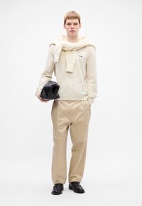 Giovane in maglione crema con sciarpa di maglia, pantaloni beige, mocassini neri, che tiene in mano una borsa nera piegata, in piedi davanti a uno sfondo bianco.