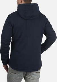 Veste bleu marine à capuche, en tissu texturé. Elle possède des manches longues et un ourlet arrondi. Détails marron sur les épaules.