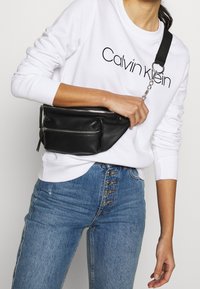 Osoba v bílé mikině Calvin Klein a modrých džínách s vysokým pasem, držící černý kožený křížový popruh tašky se zipem.