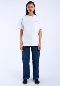 Bílý krátký rukáv tričko s zeleným textem, modré široké džíny a černé tenisky. Jednoduchý design, žádné vzory nebo další akcenty.